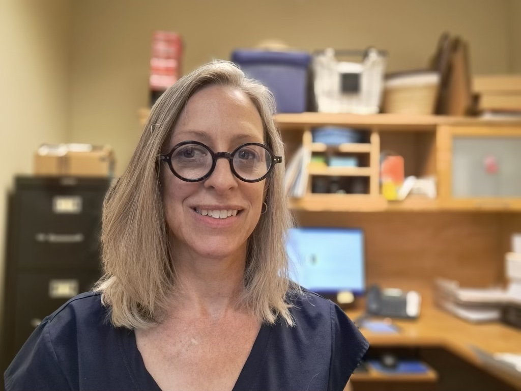 Headshot of Diane in her office, a computer screen in the background. She is wearing a navy shirt and black circular glasses